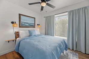 Un dormitorio con cama, ventilador de techo, un cuadro y una ventana con cortinas.