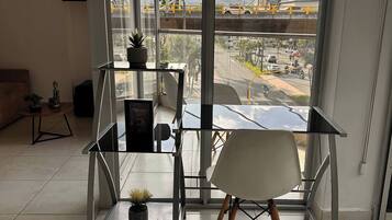 Quarto moderno com mesa e cadeira de vidro, um vaso de plantas e vista para a cidade através de amplas janelas.