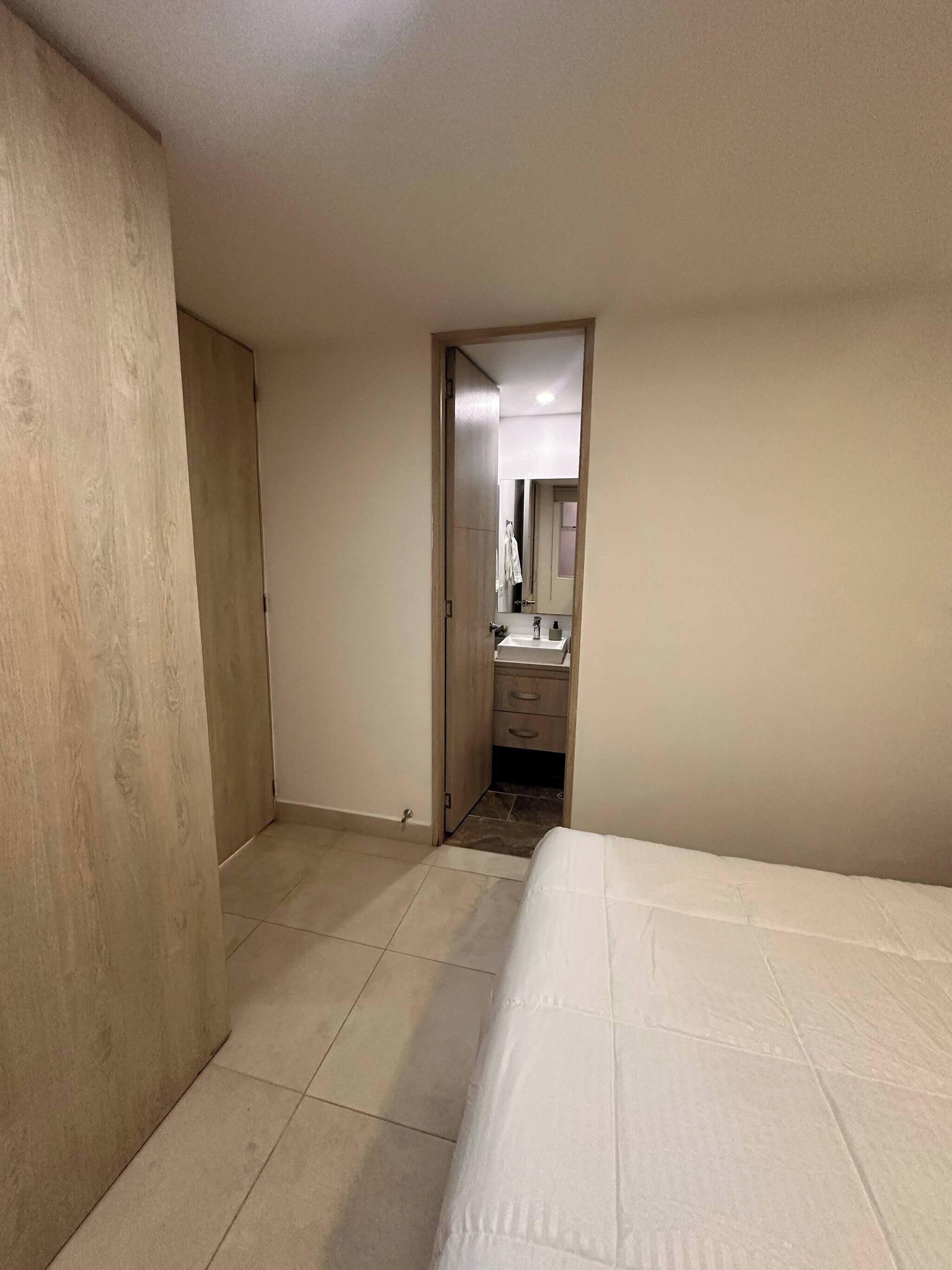 Quarto com cama, guarda-roupa de madeira, banheiro com espelho e chão ladrilhado.