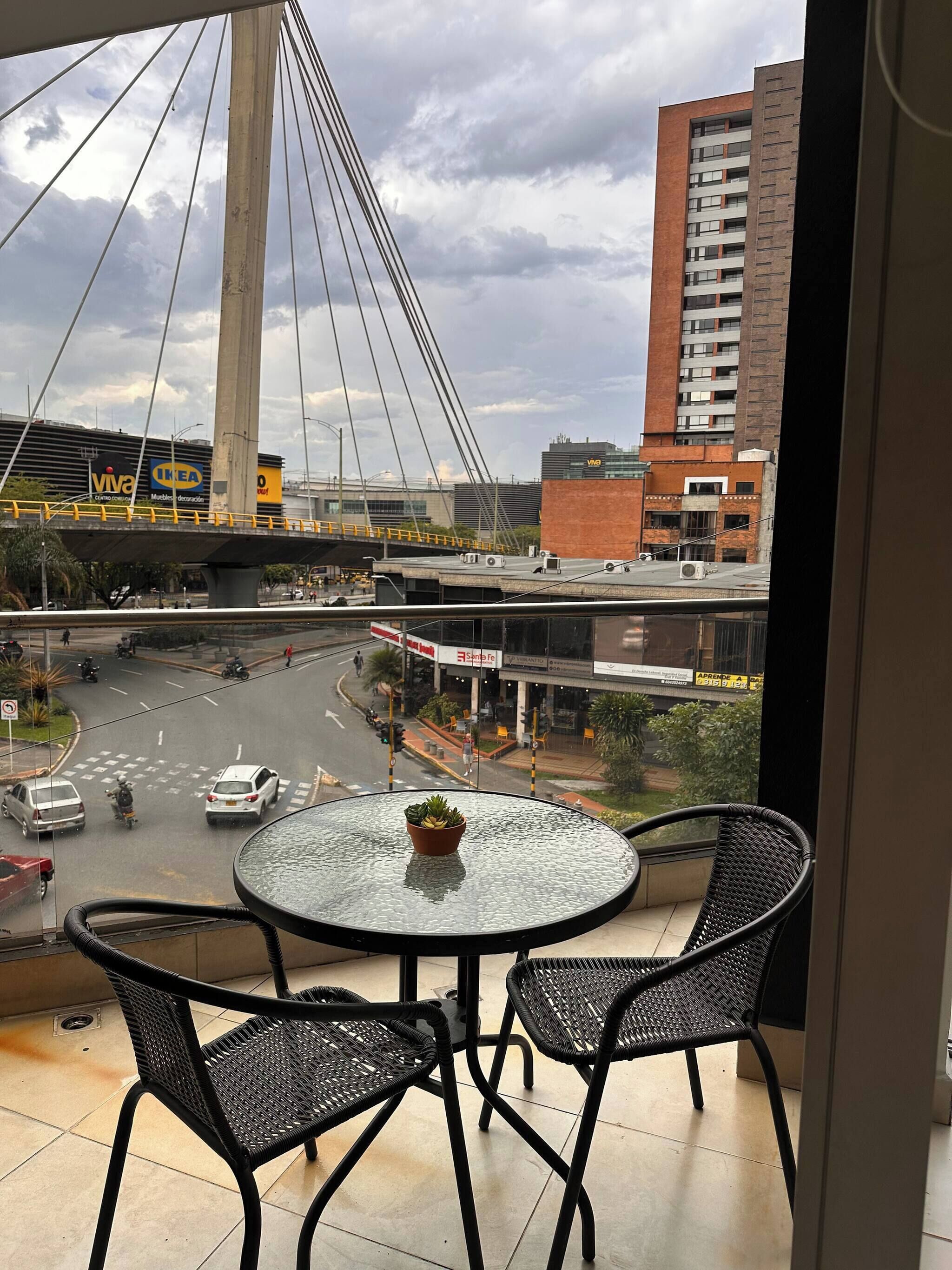 Uma varanda com mesa e cadeiras de vidro, com vista para uma rua movimentada com uma ponte e edifícios.