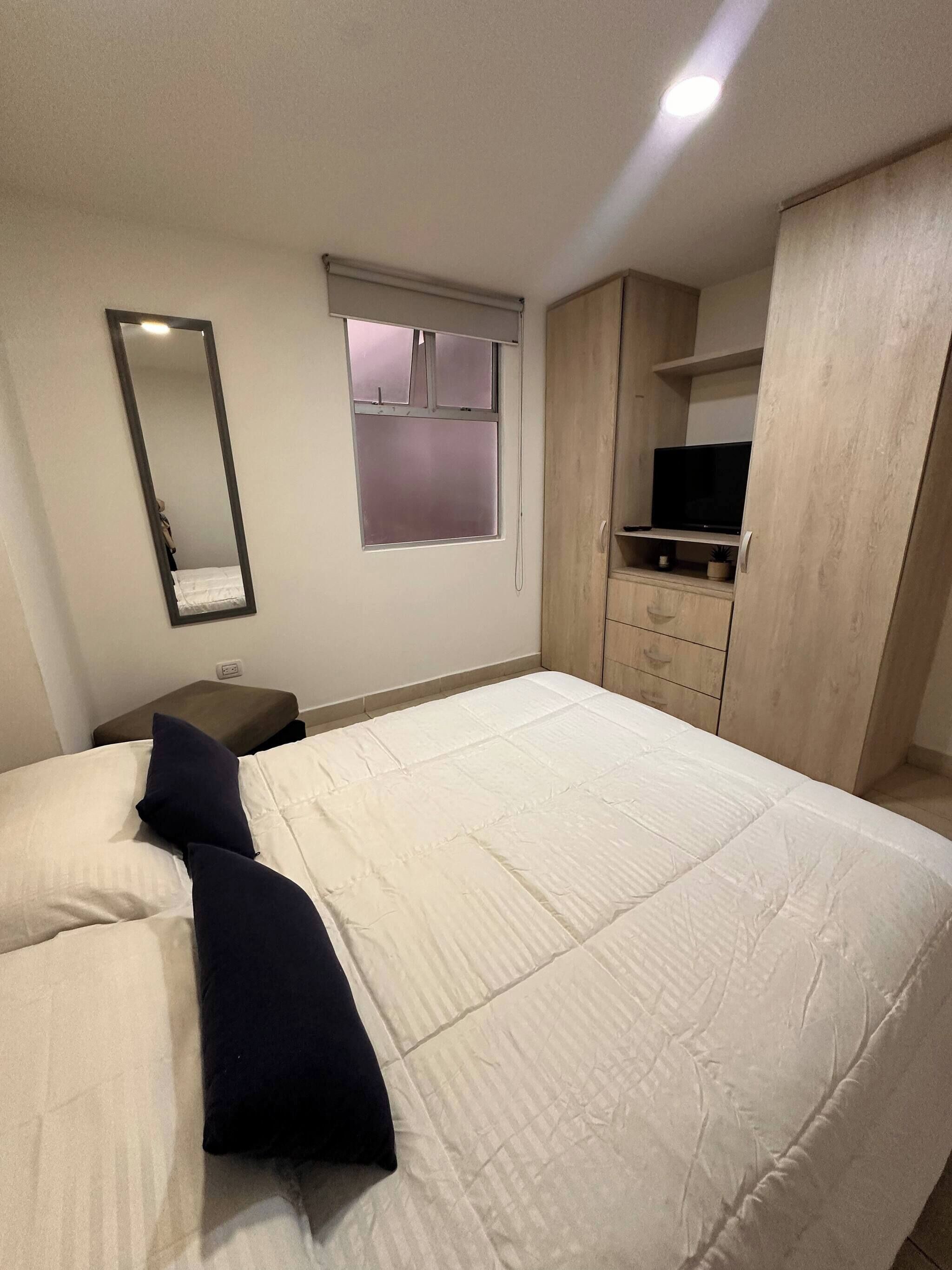 A bedroom with a bed, a bedside table, a mirror, a window, a wardrobe, and a television.