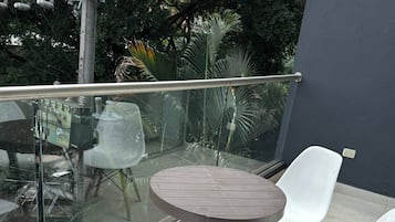 Un balcón con una mesa redonda y dos sillas blancas, barandilla de vidrio y vista a los árboles.