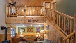 Un intérieur en bois avec une chambre à coucher située en mezzanine, un escalier et un espace de vie comprenant un canapé et des lampes.