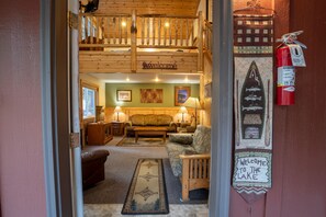 A cozy cabin interior with a wooden staircase, a welcome sign, a fire extinguisher, and a view of the living area with a couch and lamps.