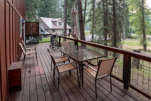 Um deck de madeira com mesa e cadeiras de vidro, com vista para uma área arborizada.