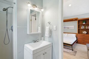 Casa de banho com chuveiro, pia e espelho. Há uma cama com cabeceira de madeira e uma estante com livros e uma planta.