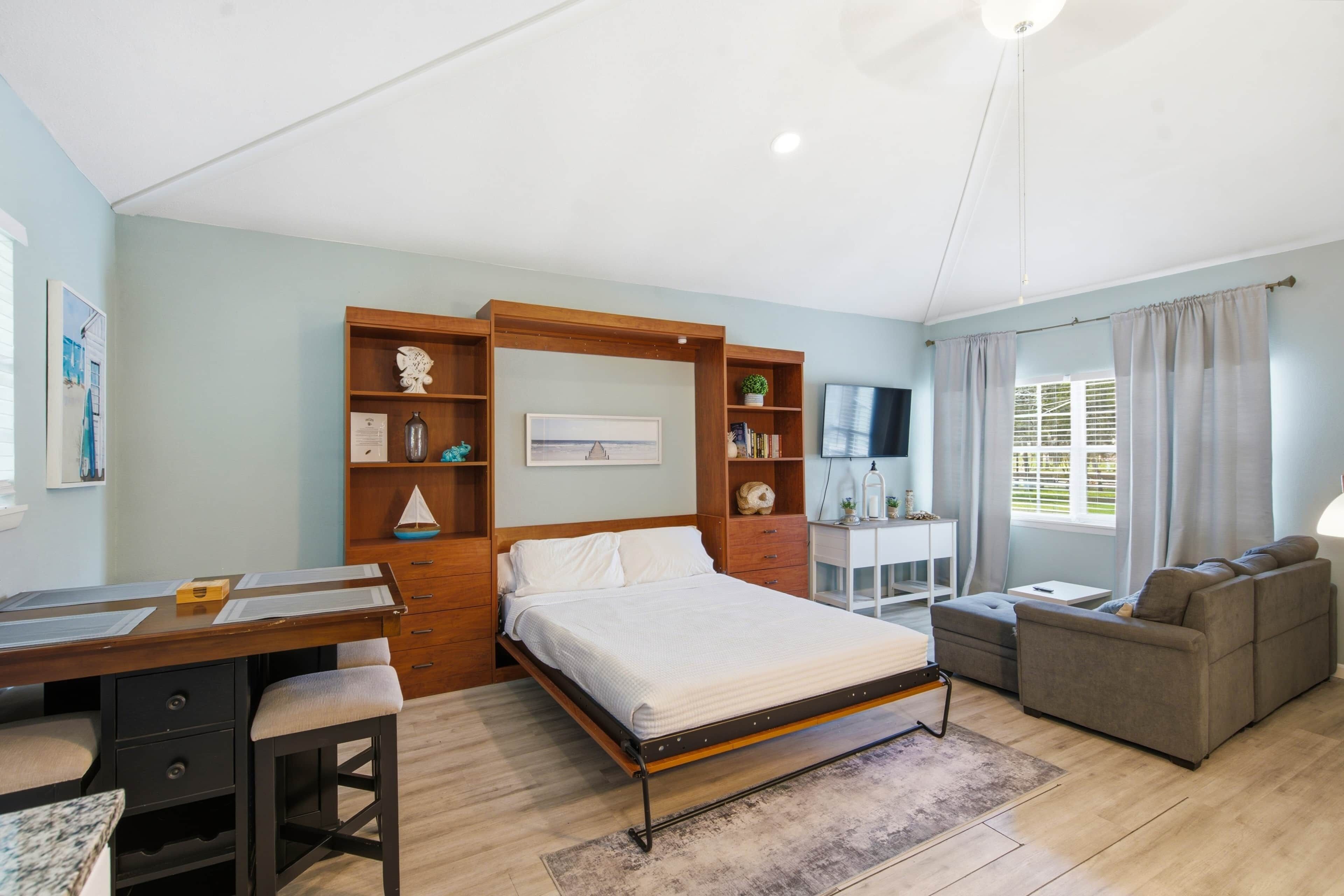 A bedroom with a bed, a desk, a chair, a TV, and a window with curtains.