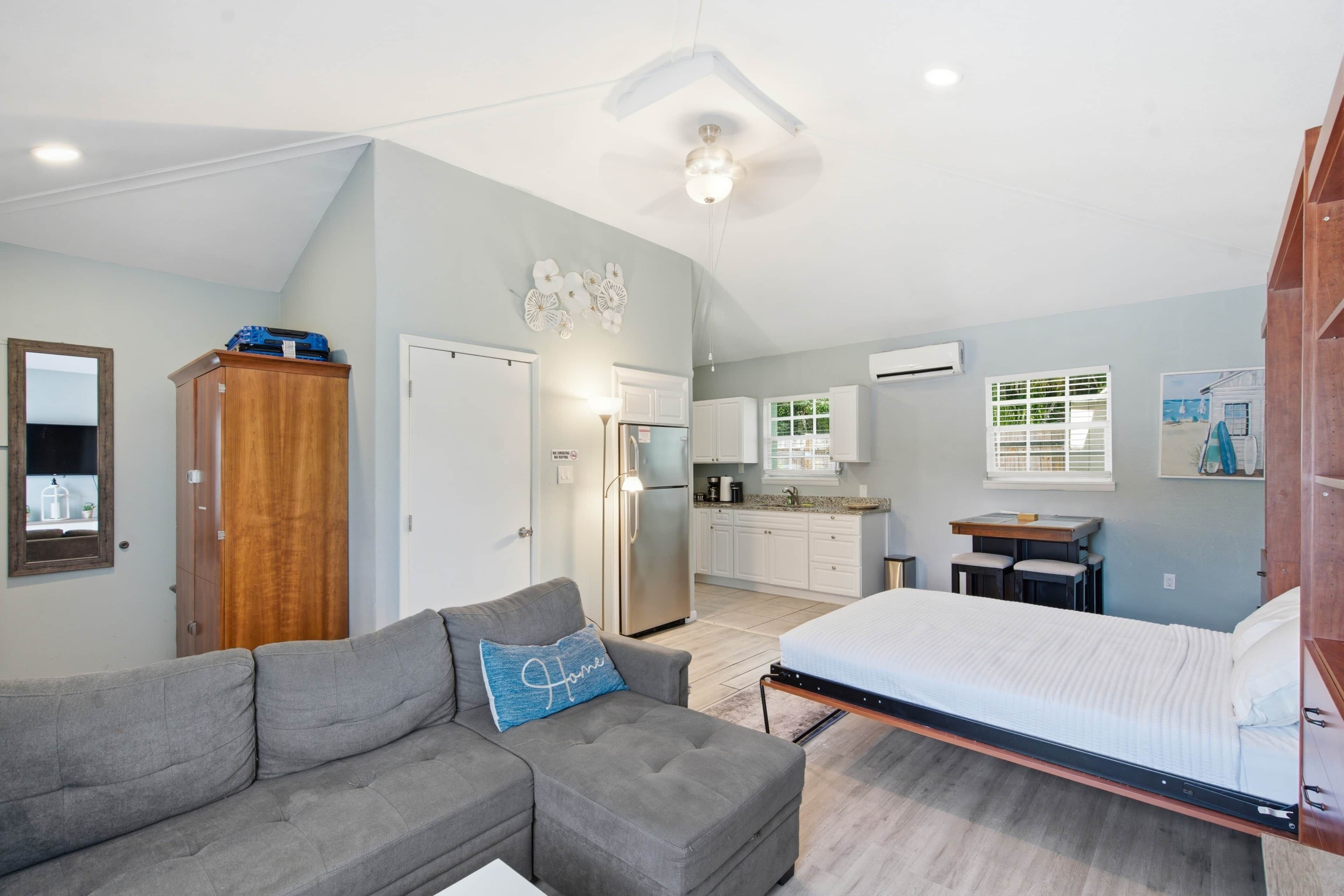 Un espacio habitable compacto con kitchenette, un sofá gris con un cojín decorativo, una cama y un armario de madera.