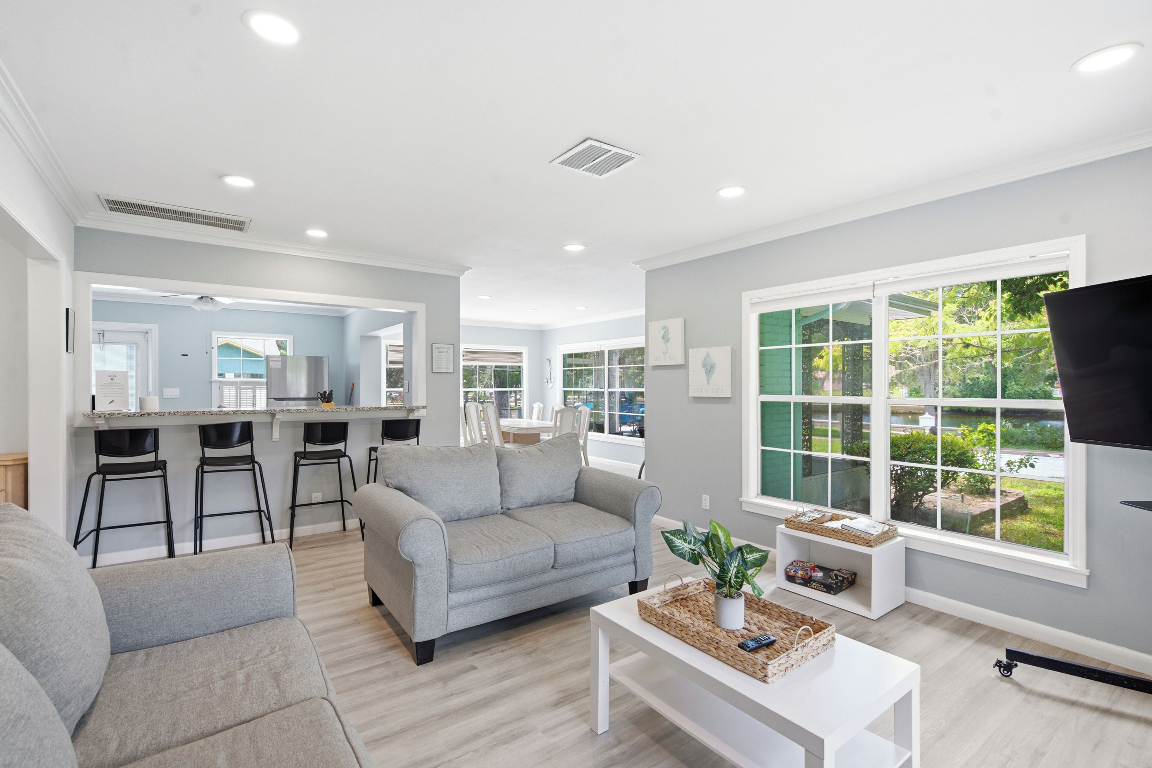 A modern living room with a grey sofa, a coffee table, and a TV. There is a kitchen area with a fridge and bar stools. The room has large windows with a view of greenery outside.