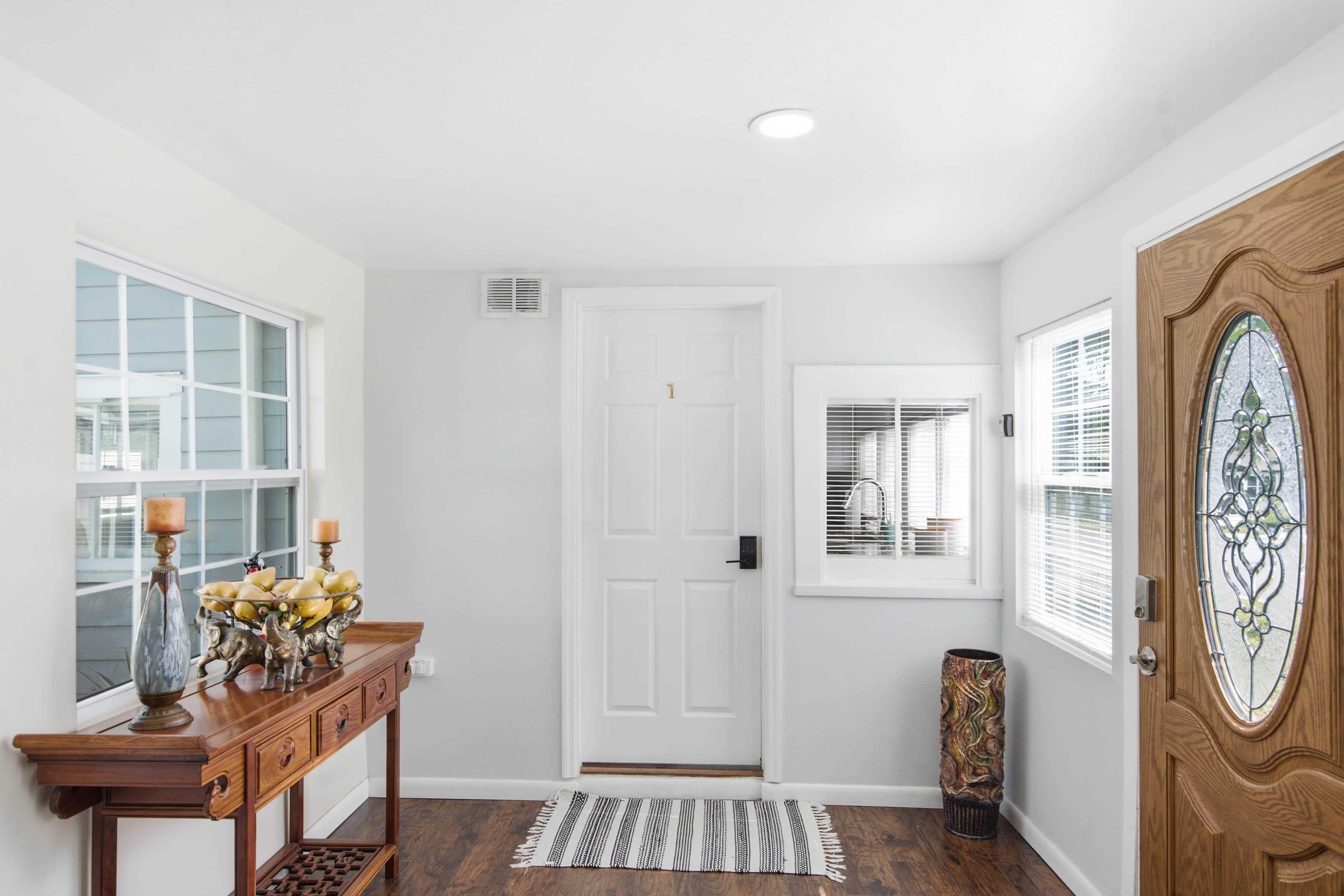 A clean, well-lit entryway with a wooden door, a white wall, a window with blinds, and a small table with decorative items.
