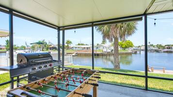 A covered outdoor area with a foosball table and a grill, overlooking a body of water with boats and houses.