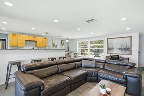 A modern living room with a leather sectional sofa, a kitchen area with wooden cabinets, and a large window with blinds. - 4 Mi to Beach! Waterfront New Port Richey Retreat (Pasco County)