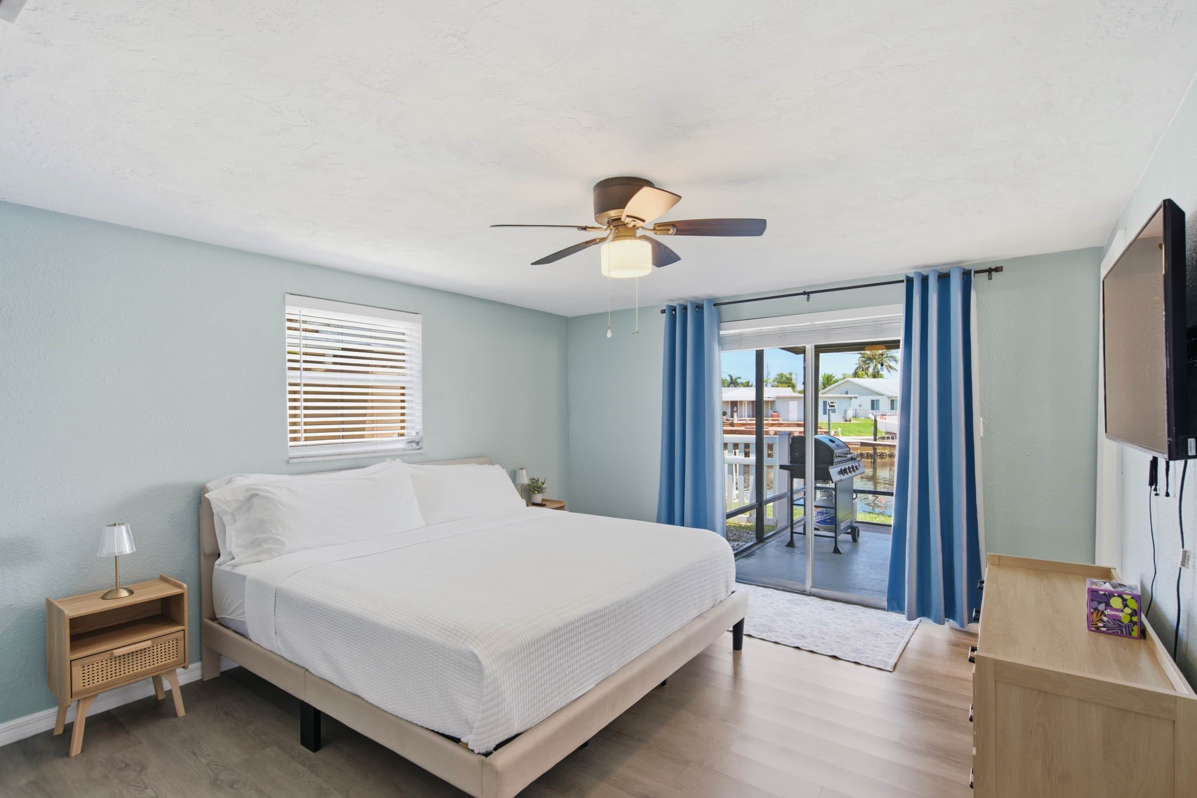 A bedroom with a bed, a ceiling fan, a window with blinds, and a view of an outdoor area.