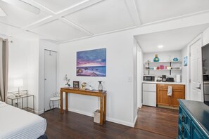 Ein kleines Hotelzimmer mit Bett, Nachttisch, Schreibtisch, Stuhl, einem Bild, einer Küchenzeile und einem Badezimmer.