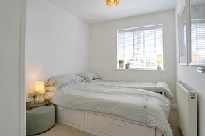 A bedroom with a bed, bedside table, lamp, window, radiator, and a small potted plant.