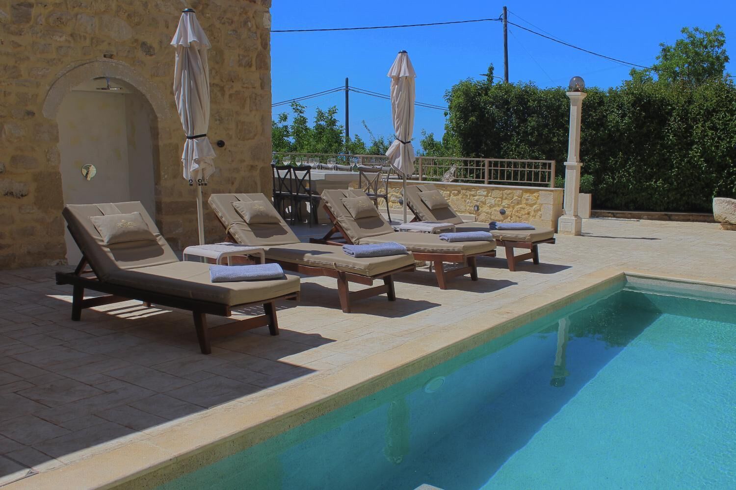 A poolside area with lounge chairs and umbrellas, a stone wall, and a clear blue sky.
