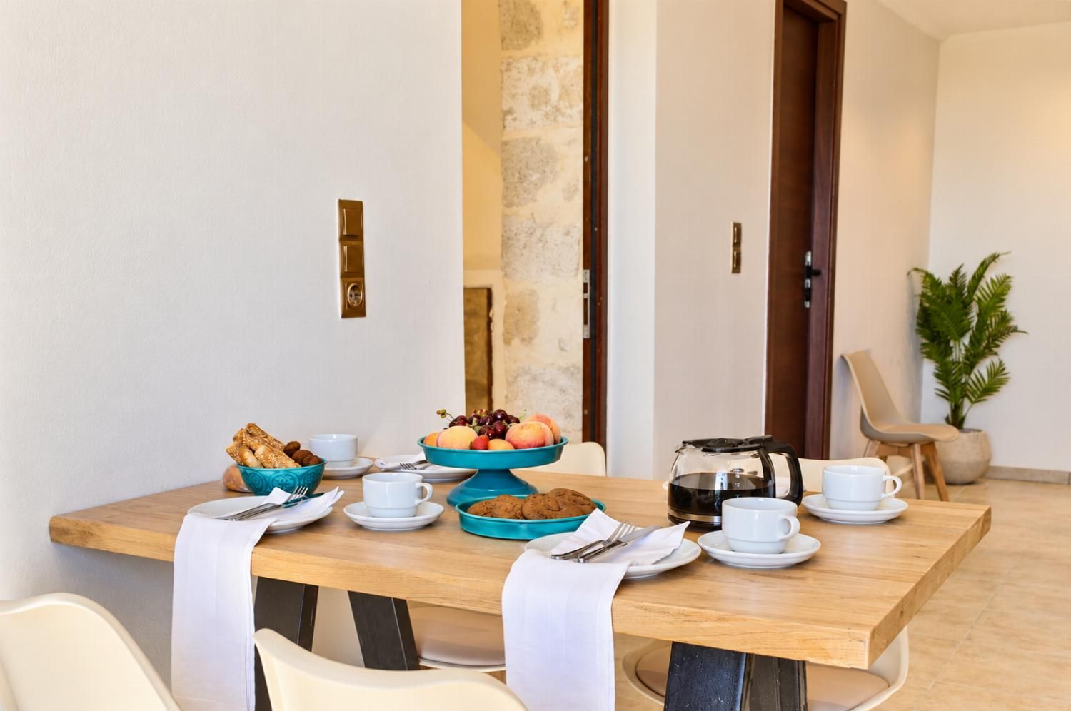 A dining table set with plates, bowls, and cups, with a coffee pot and a plant in the background.