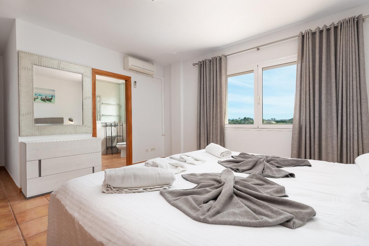 A hotel room with a bed, a large mirror, a window with curtains, and an air conditioning unit.