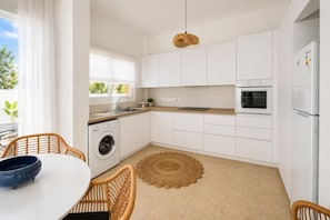 A modern kitchen with white cabinetry, a built-in oven, a washing machine, and a dining area with wicker chairs and a round table. - Villa Kori - Two Bedroom Villa, Sleeps 5 (Pernera)