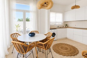 Une cuisine moderne avec des meubles blancs, une table de salle à manger ronde entourée de chaises en osier et une grande fenêtre ornée de rideaux légers.