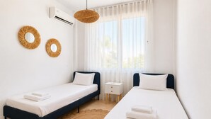 Ein Hotelzimmer mit zwei Einzelbetten, einem kleinen Nachttisch, einer wandmontierten Klimaanlage und einem großen Fenster mit durchsichtigen Vorhängen.