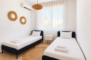 Ein Hotelzimmer mit zwei Einzelbetten, einem kleinen Nachttisch, einer wandmontierten Klimaanlage und einem großen Fenster mit durchsichtigen Vorhängen.