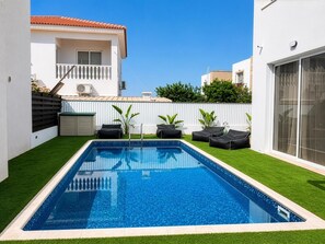 Ein rechteckiger Swimmingpool mit glasklar blauem Wasser, umgeben von künstlichem grünen Rasen, neben einem weißen Gebäude mit Balkon.