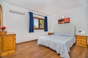 Ein Schlafzimmer mit einem Bett, einem Nachttisch, einer Kommode, einem Fenster mit Vorhängen und einer Wand-Klimaanlage.