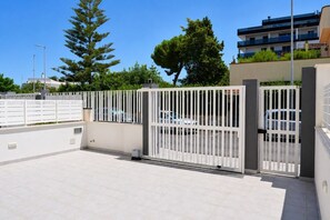 Zona d'aparcament tancada amb una tanca blanca, un cotxe a l'interior i un edifici amb balcons al fons.