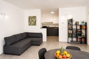 A modern living space with a sectional sofa, a dining table with fruit, a kitchen in the background, and a bookshelf filled with books and decorative items. - GuestHost - Comfy Verandah Flat X6 Near the Sea (Trani)