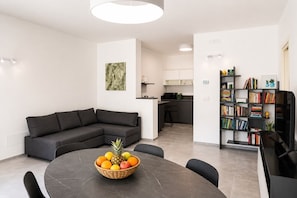 Ein modernes Wohnzimmer mit einem dunkelgrauen Ecksofa, einem runden Esstisch mit Obstschale, einer Küchenzeile und einem Bücherregal mit Büchern und Dekoration.