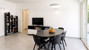 A modern dining area with a round table, black chairs, a fruit bowl, a black TV, and a bookshelf. - GuestHost - Comfy Verandah Flat X6 Near the Sea (Trani)