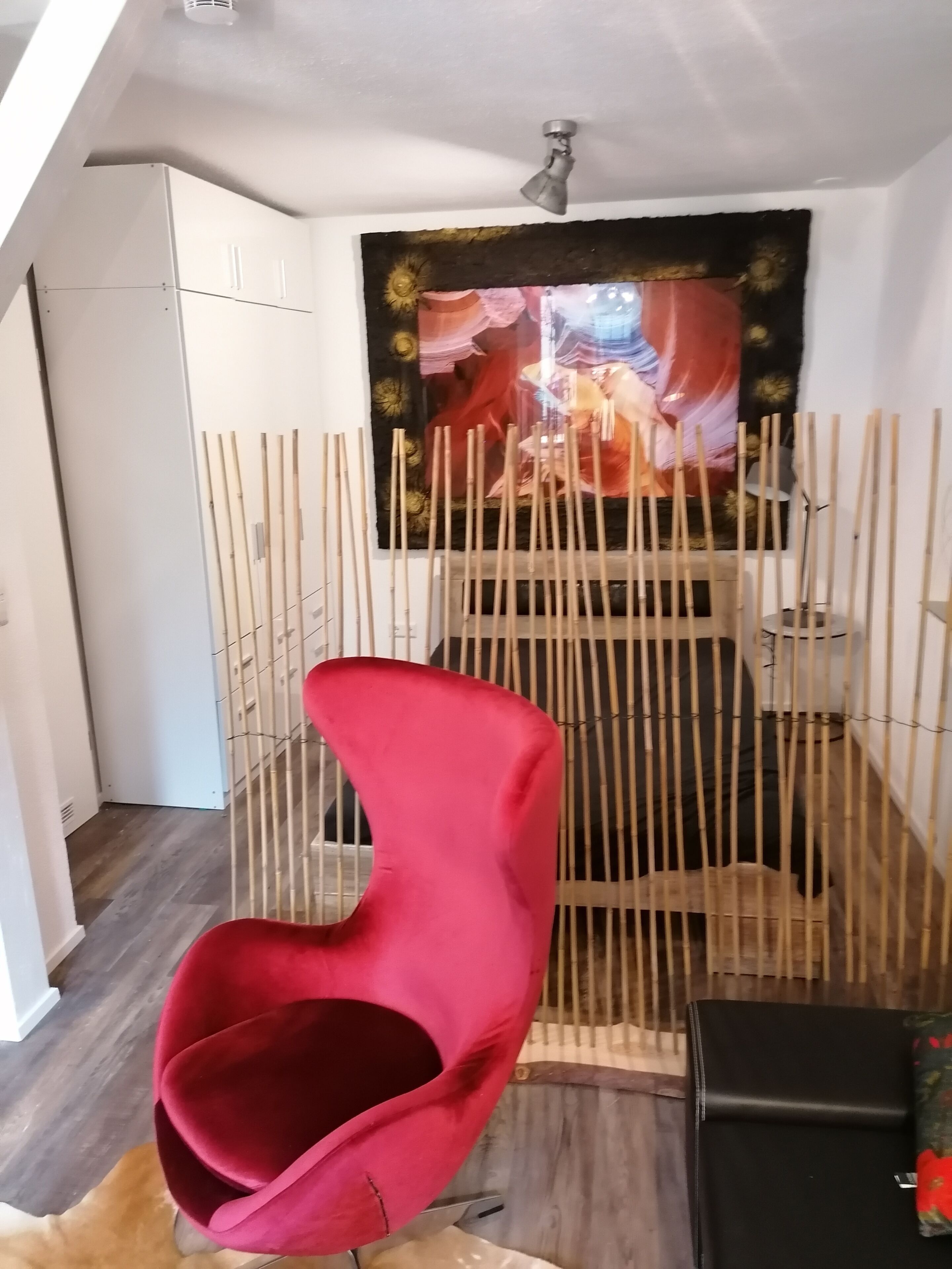 Ein Zimmer mit einem roten Sessel, einem Bett mit Kopfteil, einer Wandteppich und einem Deckenventilator.