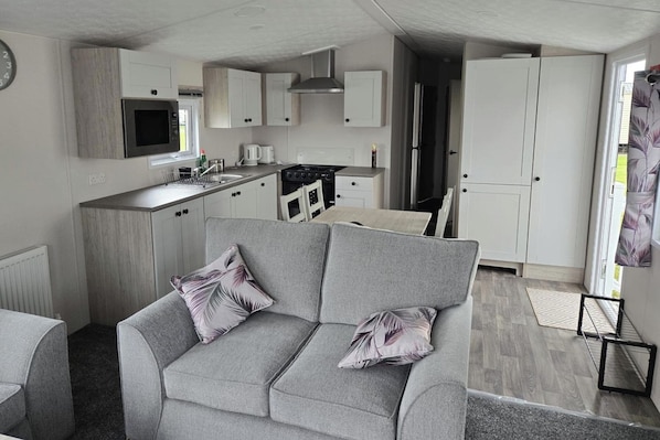 Un espacio habitable compacto con kitchenette, sofá gris con cojines estampados y vistas al exterior.