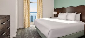 Ein Hotelzimmer mit einem großen Bett, einem Nachttisch mit Lampe, einem Schminktisch und Blick auf den Ozean durch ein mit Vorhängen drapiertes Fenster.
