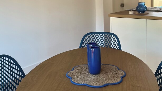 Ein runder Holztisch mit einer blauen Vase, umgeben von blauen Stühlen und einer gewebten Tischmatte.