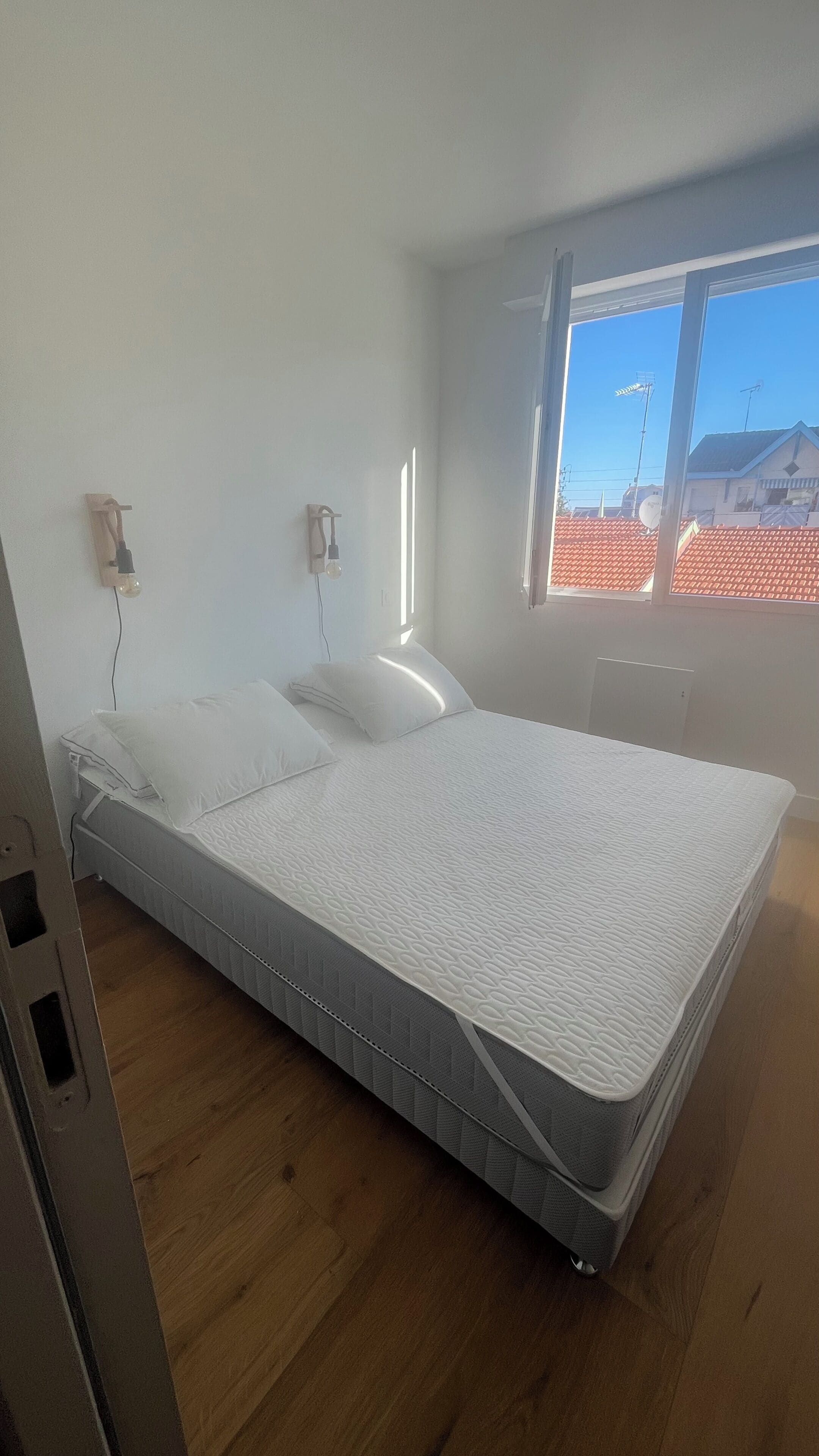 Une chambre avec un lit simple, une fenêtre donnant sur des immeubles et un luminaire fixé au mur.