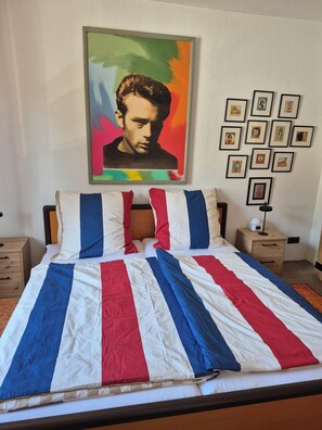 Ein Bett mit gestreifter Bettdecke, eine Wand mit einem Porträt und gerahmten Bildern sowie ein Nachttisch mit einer Lampe.
