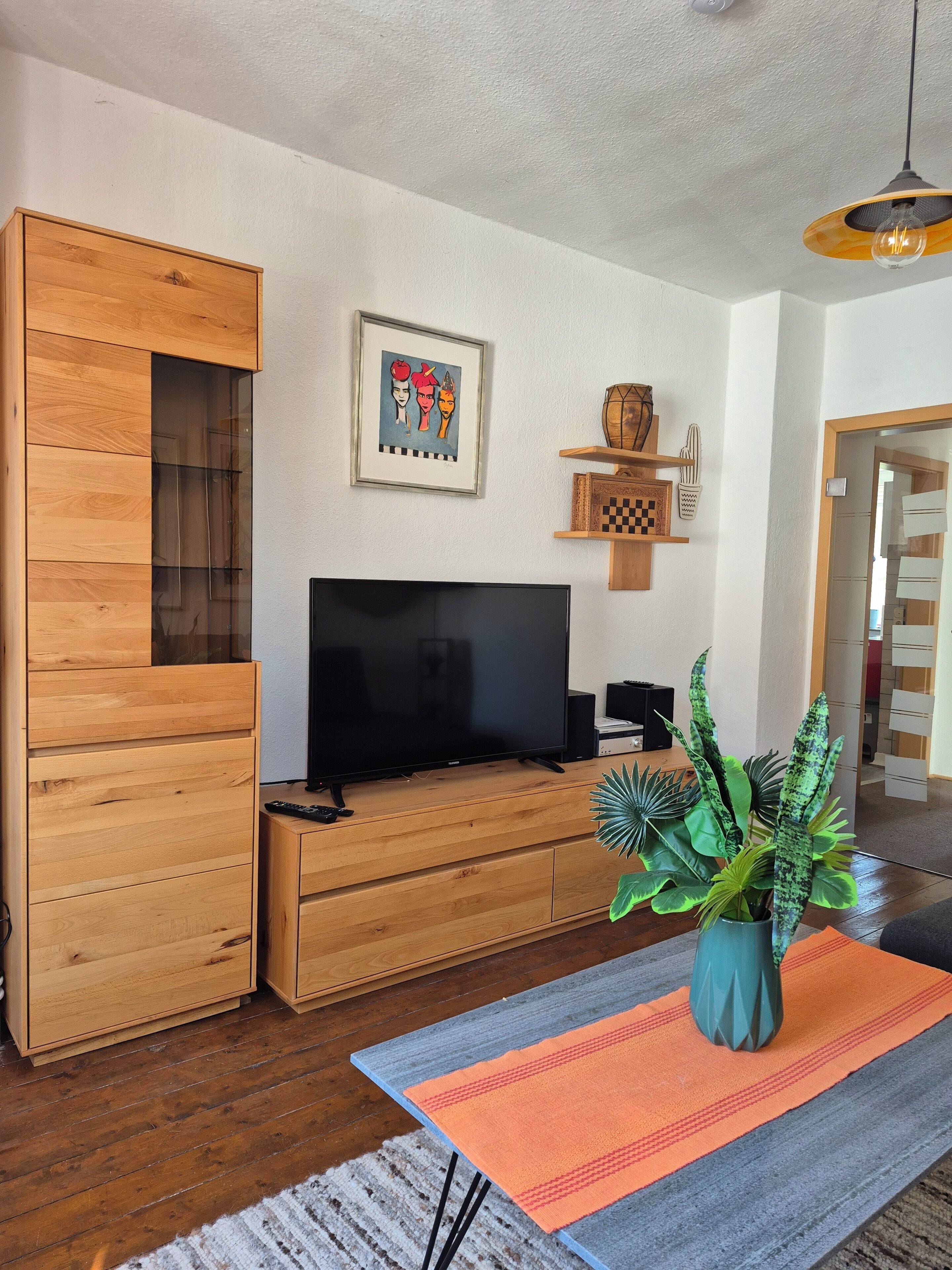 Una sala de estar moderna con un mueble de entretenimiento de madera, un televisor de pantalla plana, una obra de arte enmarcada y una planta en maceta.
