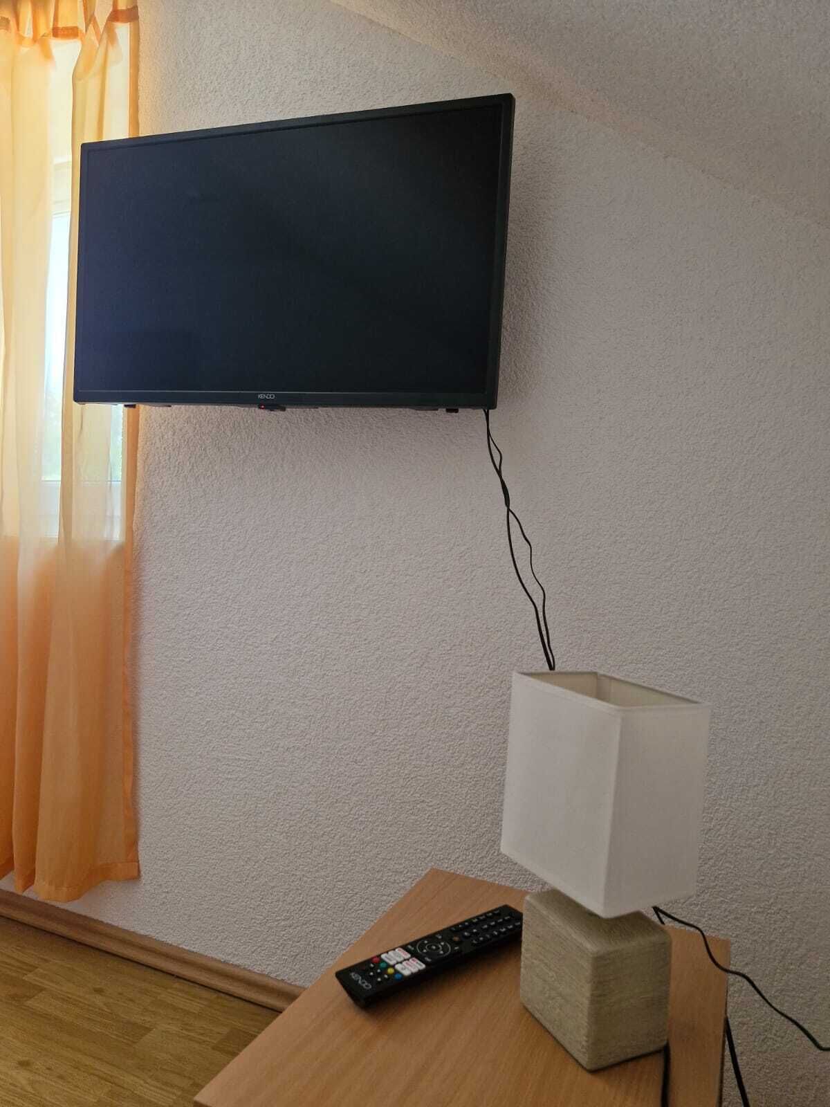 Et fjernsyn monteret på en væg, en fjernbetjening på et træbord, og en lampe med hvide skærm.