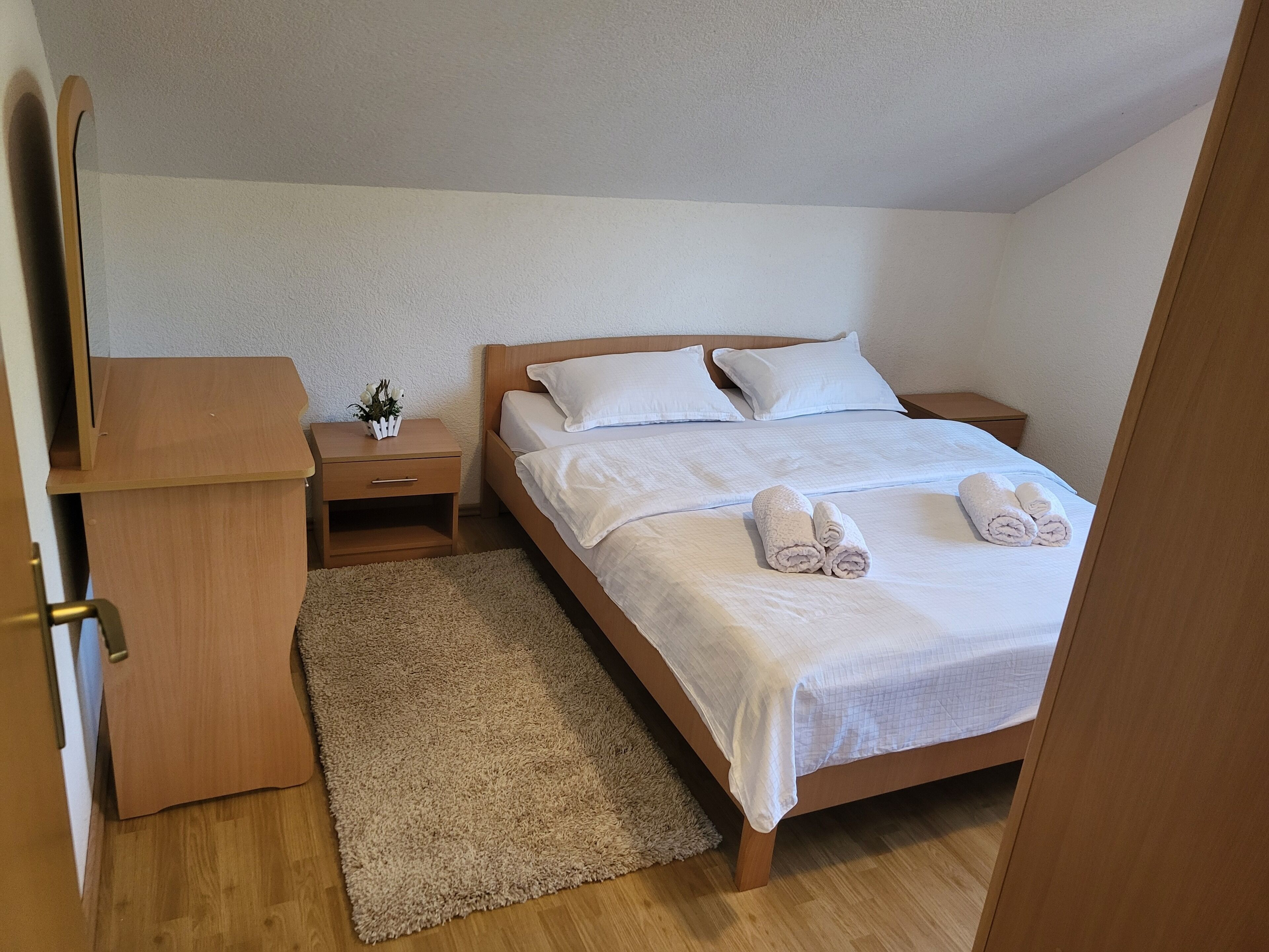 A bedroom with a wooden bed, white linens, a bedside table with a plant, and a wooden dresser.