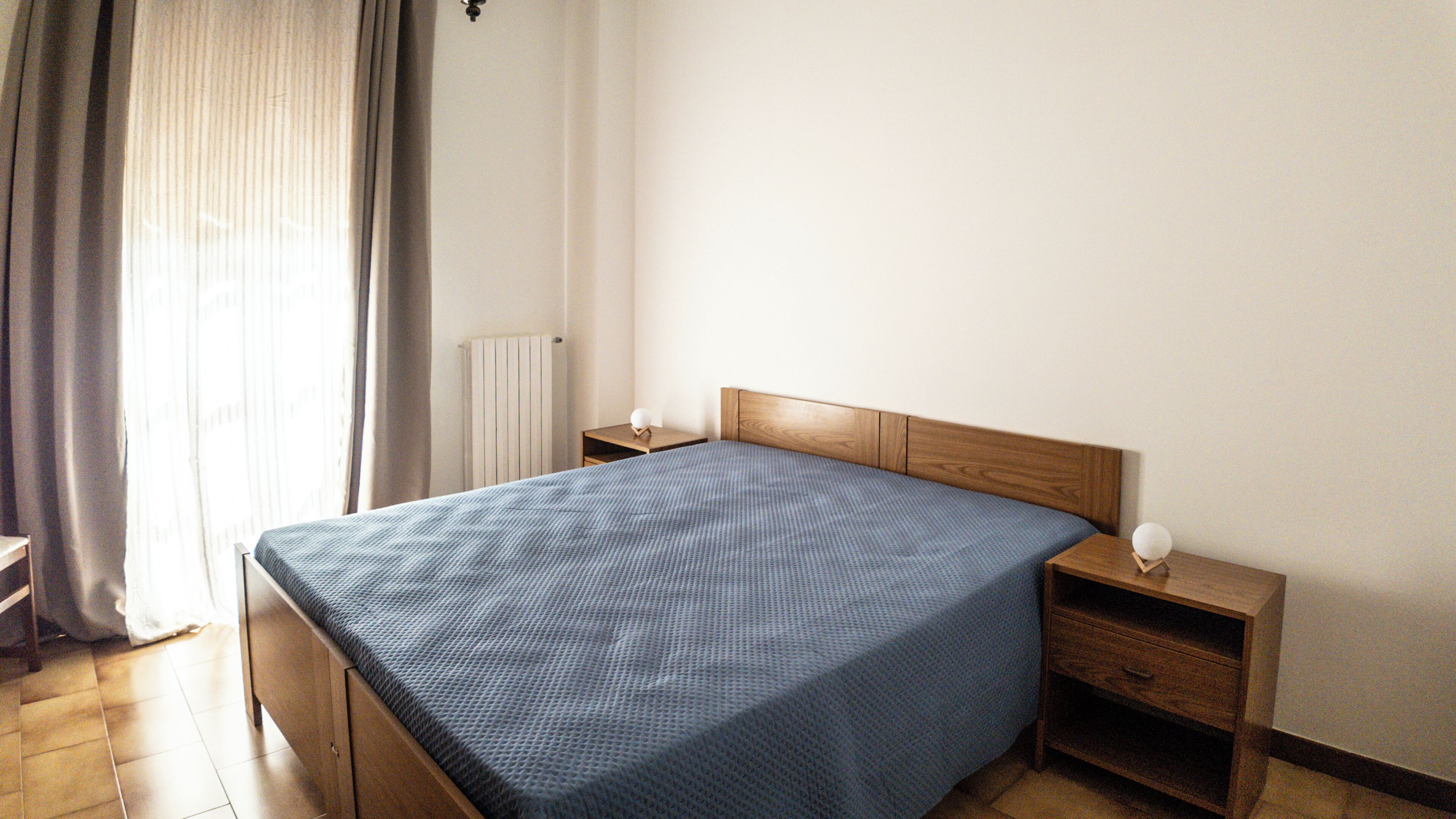 Un dormitorio con cama, mesita de noche, cajón, radiador y ventana con cortinas.