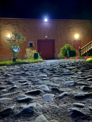 A cobblestone pathway leading to a building with a red door, a staircase, and a small tree with colorful lights.