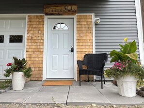 A front door with a wooden sign above it, a wicker chair, and two potted plants on the porch. - Charming 3-bedroom house in fantastic Bar Harbor w/ AC, art, private studio (Bar Harbor)