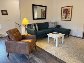 A living room with a brown leather chair, a green sofa, a white coffee table, and a floor lamp. - Charming 3-bedroom house in fantastic Bar Harbor w/ AC, art, private studio (Bar Harbor)