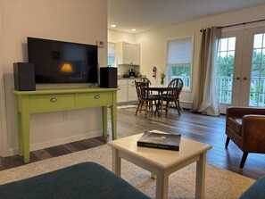 Een woonkamer met een televisie, een tafeltje met een boek, een bank en een eethoek met stoelen.