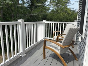 Eine Holzterrasse mit weißer Reling, einem Korbstuhl und einem weißen Gebäude im Hintergrund.