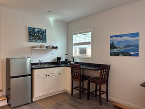 A compact kitchen with white cabinets, a built-in refrigerator, a black countertop, and two wooden chairs. - Charming 3-bedroom house in fantastic Bar Harbor w/ AC, art, private studio (Bar Harbor)