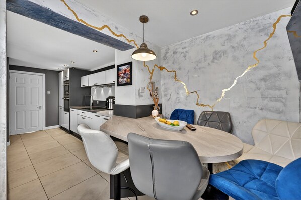 A modern dining area with a marble wall, a wooden table, and a mix of grey and blue upholstered chairs. - Charming Devon Cottage With Garden (West Kilbride)