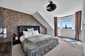 Un dormitorio con cama, mesita de noche, lámpara, ventana con cortinas y vista al mar.